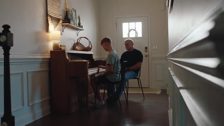 Young man plays the piano while a man beside him watches closely