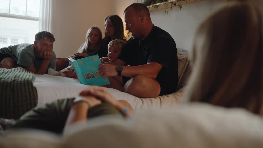 A large family gathered in a bedroom around the father reading to the youngest boy from a childrens book