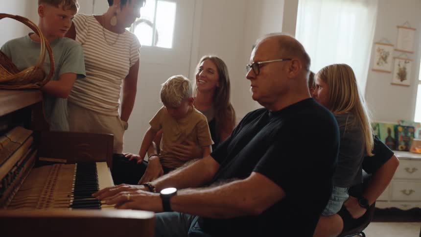 Man plays the piano while family members watch and interact around him