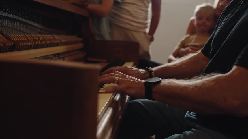 A man plays the piano as his family watches nearby indoors