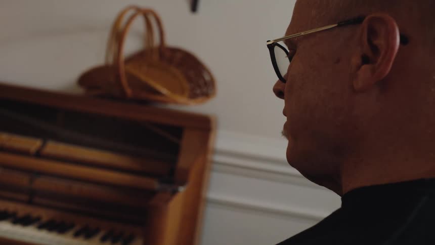 A man wearing glasses looks toward an upright piano with its front panel removed exposing the keys and hammers
