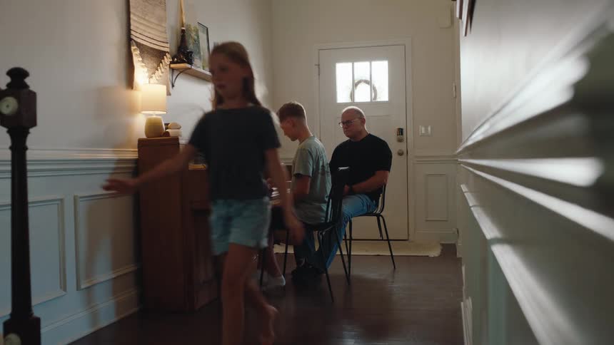 A boy plays a piano while a man sits beside him and watches and a girl walks past them in a hallway