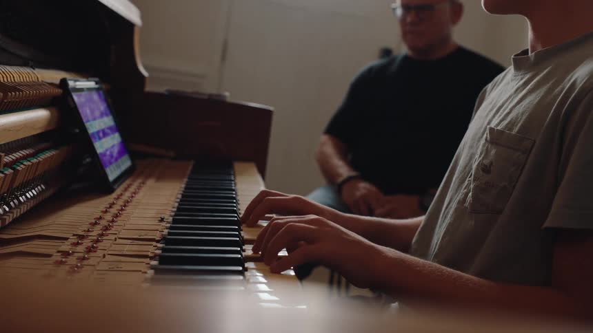 An older man teaches a teenager piano using a tablet indoors