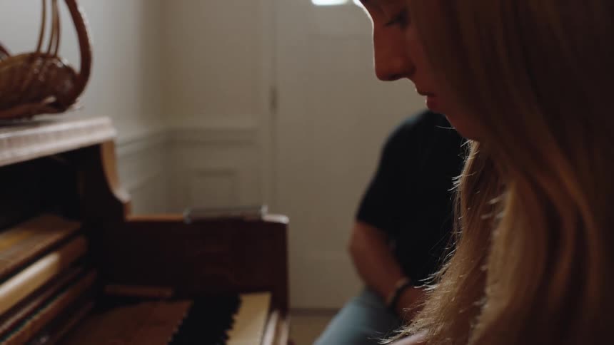 A young woman is playing a song on an upright piano in a bright room with her fingers pressing the keys while a man sits nearby in the background
