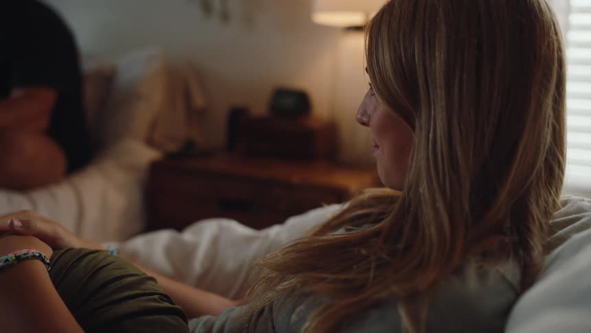 Young woman with long brown hair sitting on a chair inside a bedroom smiling warmly a lamp glows softly at her side while friends are present in the room including a young man sitting on the bed beside her