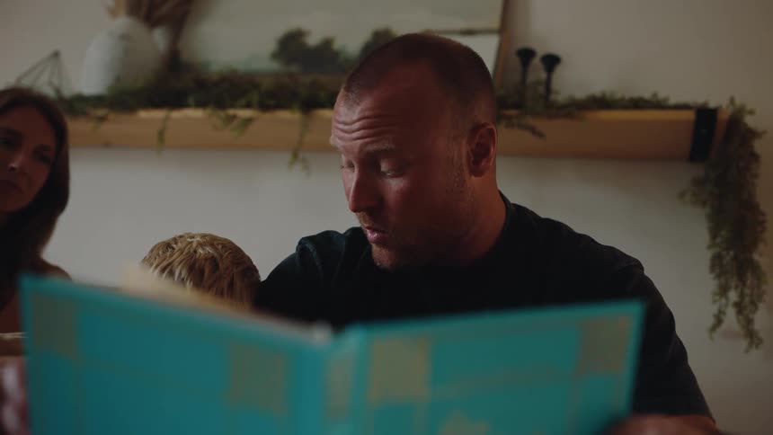 A man is sitting at a kitchen table and reading a book aloud to a little child with a woman sitting next to them in the cozy interior of a house