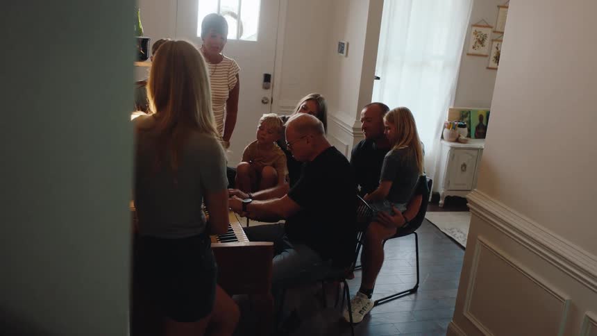 A family gathers around a man playing the piano in a living room