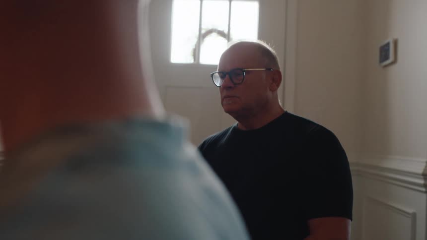 An older man wearing glasses and a black shirt sits indoors looking forward talking and nodding his head while another person is seen nearby