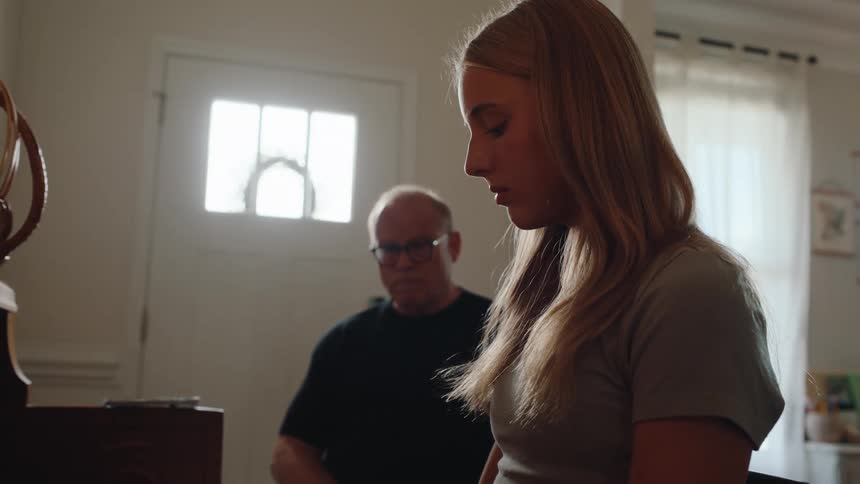 A side profile shot shows a young blonde woman with her eyes downcast sitting in what appears to be a living room an older man sits behind her blurred and out of focus