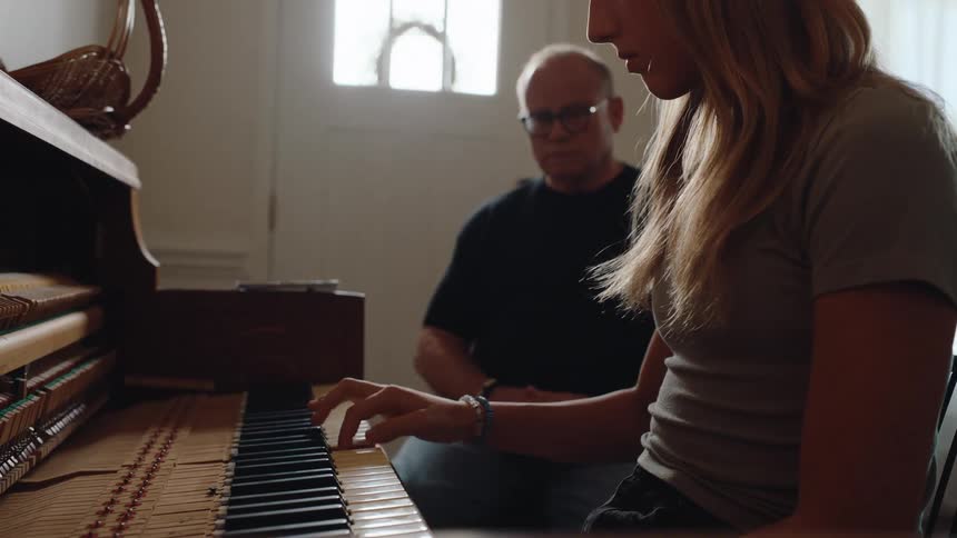 A female pianist is playing a song on a piano with a man in the background with the camera focused on the piano keys and her fingers