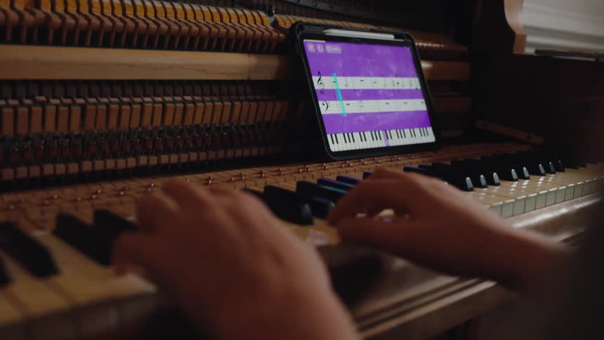 A pianists hands play a song on an upright piano while a tablet with a purple music teaching app and a digital keyboard rests on the keys