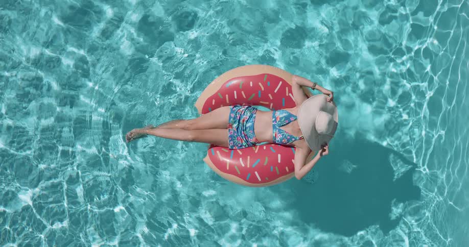 A woman in a blue patterned swimsuit and a white sun hat relaxes on a pink donut shaped inflatable in a shimmering blue swimming pool