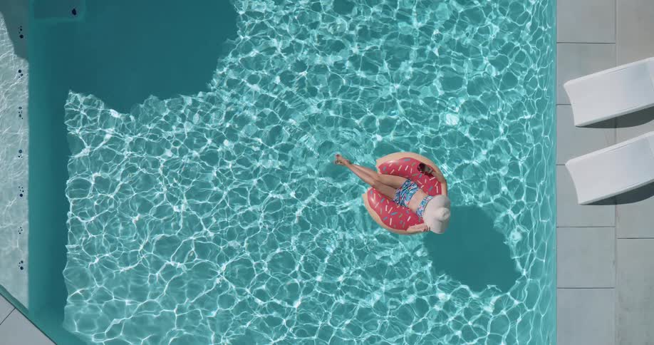 A woman wearing a sun hat is floating on a pink donutshaped inner tube in a clear blue swimming pool on a sunny day