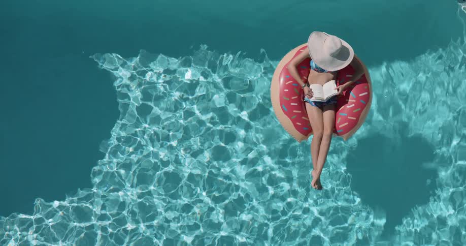 A woman wearing a sun hat floats on a donut shaped inflatable in a swimming pool reading a book