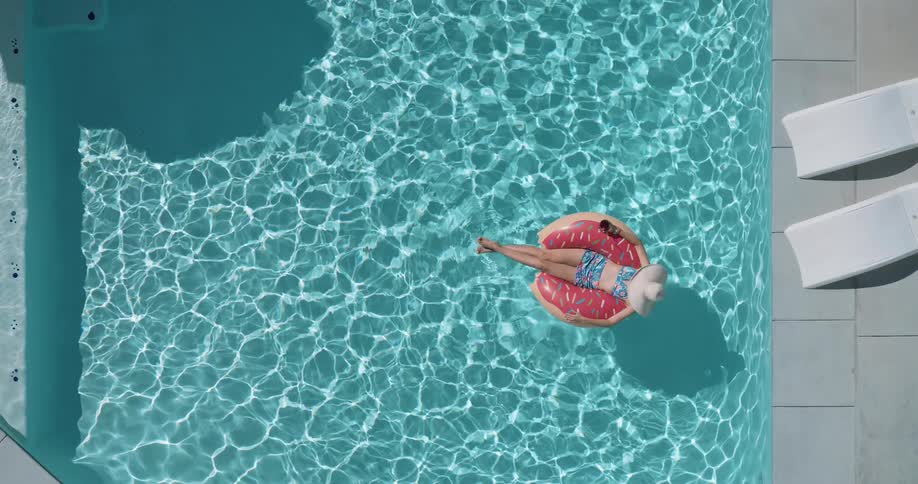 A woman wearing a sun hat is floating on a pink donutshaped inner tube in a clear blue swimming pool on a sunny day