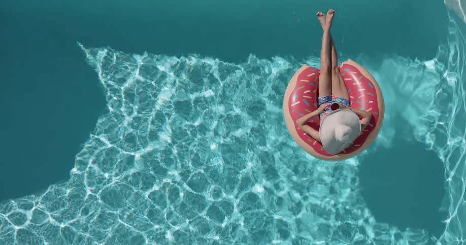 A woman wearing a sun hat relaxes in a pool floating on a doughnut shaped buoy as she sips red wine from a glass