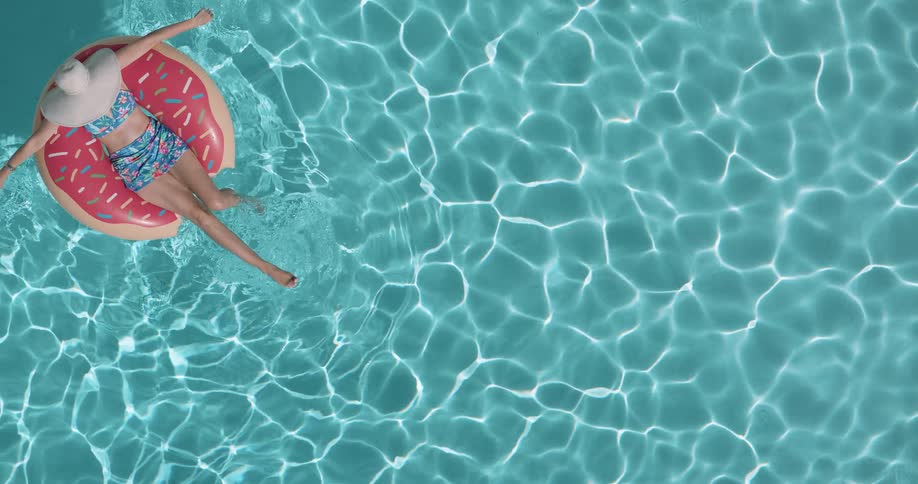 An overhead shot shows a woman in a sun hat and a twopiece bathing suit relaxing on a donutshaped pool float she drifts slowly across a sparkling blue swimming pool