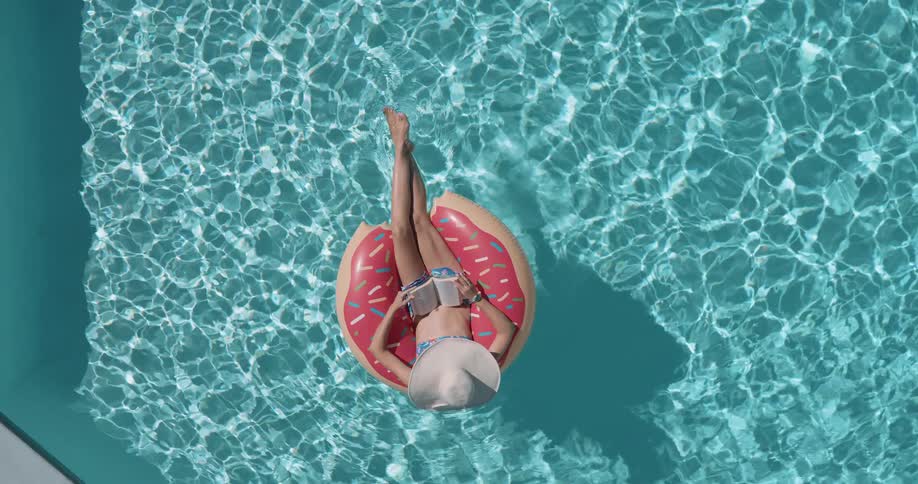 A topdown aerial view shows a woman in a sun hat reading a book while relaxing on a donutshaped pool float in a sparkling blue swimming pool