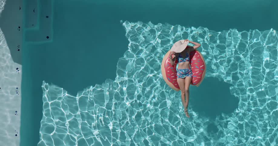 Woman wearing a hat and swimsuit lying on a ring float in a swimming pool relaxing under the bright sun on a clear day
