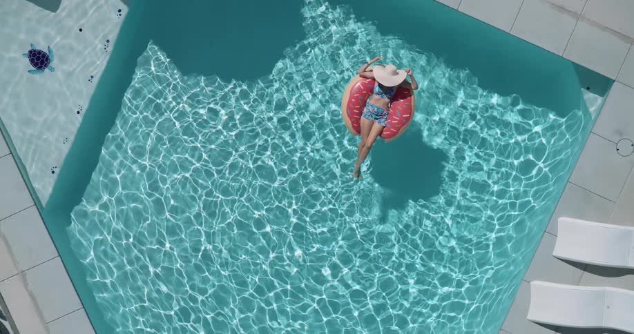 A woman wearing a sun hat is floating on a pink donutshaped inner tube in a clear blue swimming pool on a sunny day