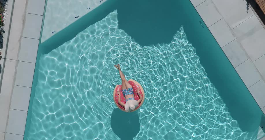 A person in a sun hat is floating on a pink donutshaped inner tube in a clear blue swimming pool on a sunny day