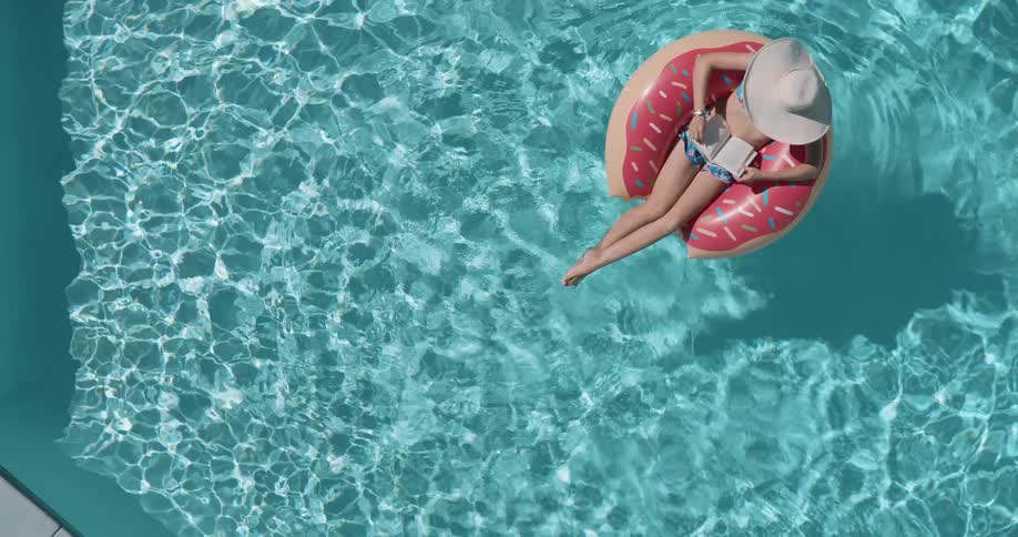An overhead shot shows a woman with a widebrimmed hat floating in a pool she is lounging on a donutshaped inner tube while reading a book on a bright sunny day the water around her is clear and a vibrant shade of blue