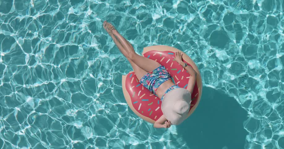A topdown aerial view shows a woman in a sun hat relaxing on a donutshaped pool float in a sparkling blue swimming pool