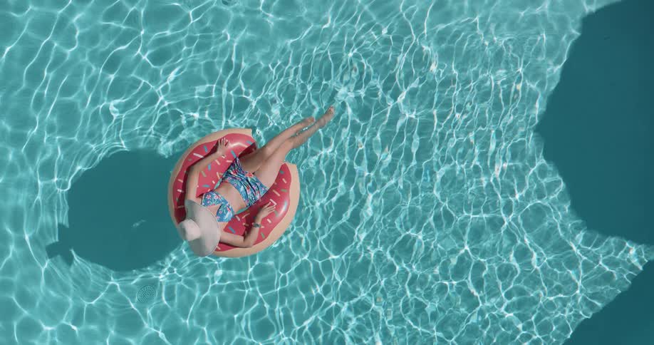 A woman in a bikini and sunglasses floats on a donut shaped inflatable in a sparkling blue swimming pool