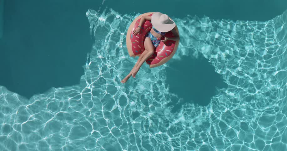 An overhead view shows a woman on a donutshaped floatie with her legs dangling in the water the sunlight creates a ripple pattern on the turquoise water of the pool as she floats gently
