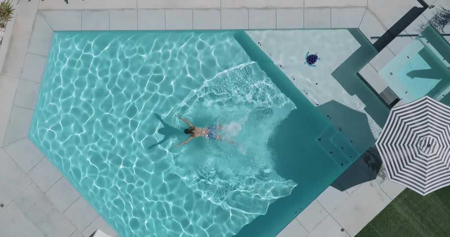 A woman wearing a swimsuit is swimming in a blue geometricshaped swimming pool on a sunny day