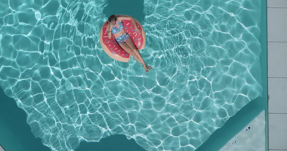 A woman wearing a blue patterned bikini and sunglasses floats on a pink donut shaped inflatable in a shimmering blue swimming pool