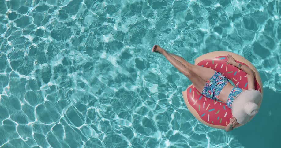 A woman wearing a sun hat is floating on a pink donutshaped inner tube in a clear blue swimming pool on a sunny day