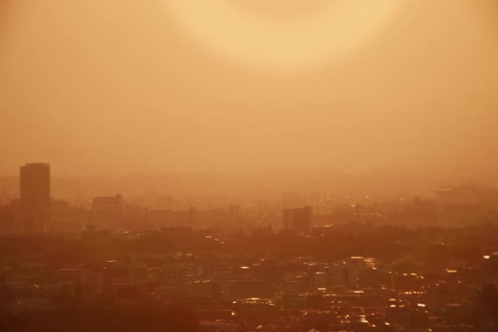 The sun sets over a hazy cityscape casting a warm orange glow across the sky and buildings