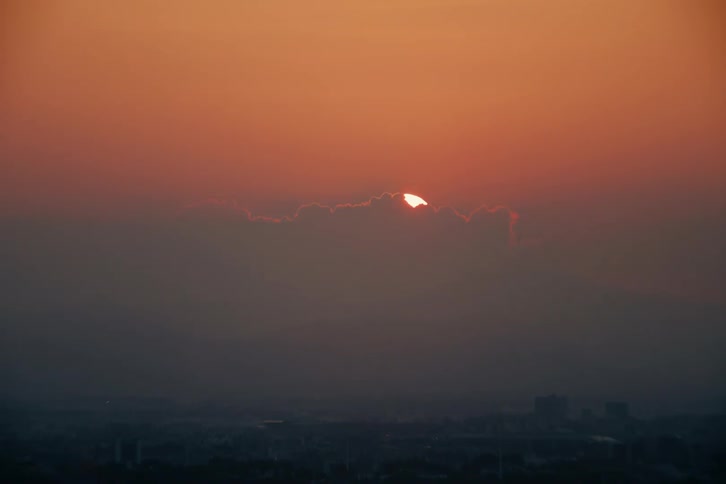 The sun dips below a line of clouds casting an orange glow across a hazy cityscape