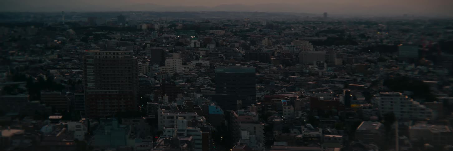An aerial shot captures a densely packed city at dusk with buildings stretching across the landscape and distant hills in the hazy background