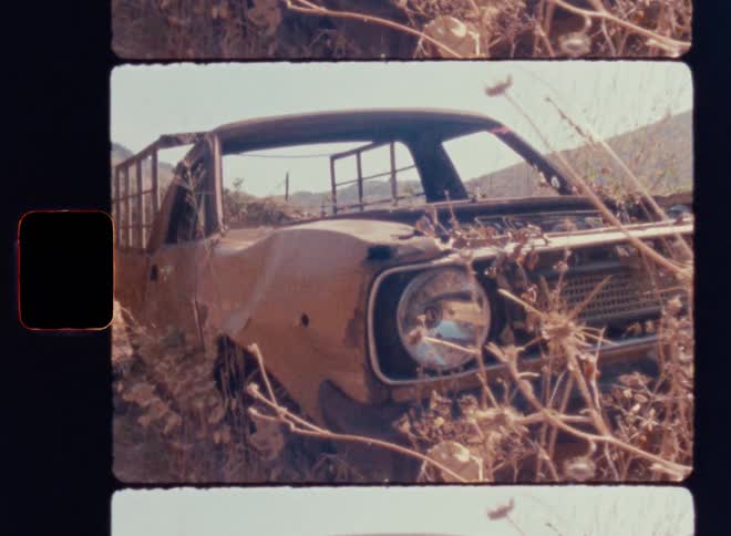 Cinematic Super 8mm close-up of a rusted abandoned vintage pickup truck overgrown with dry weeds and brush on a sun-bleached hillside, showcasing decayed metal, broken headlight and weathered frame in a handheld nostalgic film look