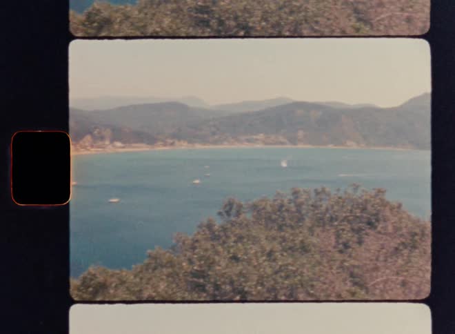Cinematic Super 8mm coastal panorama: camera pans from a sandy beach across calm turquoise water toward distant mountains, capturing a nostalgic vintage travel aesthetic with warm film grain and dreamy ocean scenery