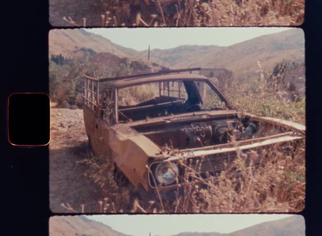 Cinematic Super 8mm shot of an abandoned rusty pickup truck overgrown with dry grass in a remote mountain landscape, featuring vintage film grain, faded colors, and a rugged rural setting with derelict structures in the background