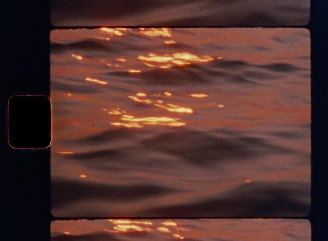 Cinematic Super-8mm close-up of gentle ocean waves reflecting warm golden and red sunset light, with shimmering highlights dancing across the water surface, captured in authentic vintage film grain and sprocket-hole framing for a nostalgic aesthetic
