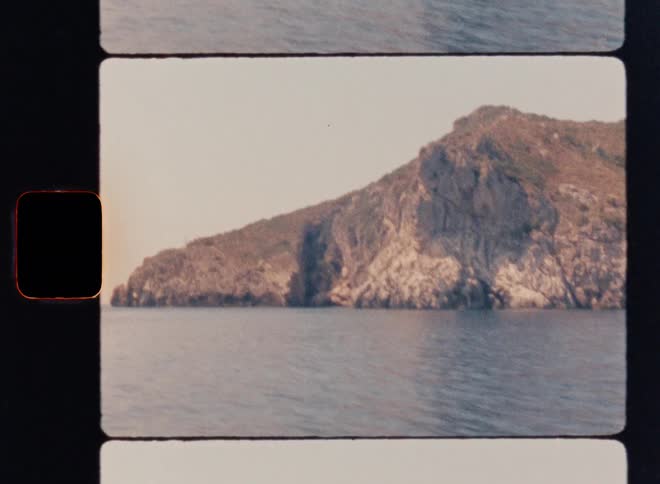 Super 8mm coastal tracking shot capturing rugged Mediterranean sea cliffs, sunlit rock formations, sparse vegetation and calm blue water as the camera moves parallel along the shoreline, framed with vintage film grain and sprocket borders