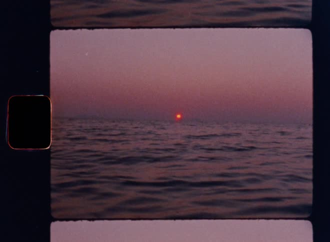 Cinematic Super-8mm shot of a deep red sun rising over calm open ocean waters, casting soft pink and purple hues across the hazy sky and gentle waves, captured with vintage film grain and visible sprocket-hole frame