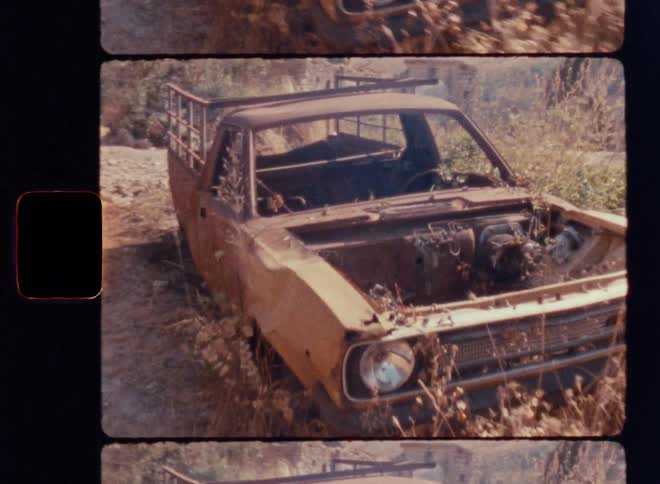 Cinematic Super 8mm shot of a rusted abandoned vintage pickup truck overtaken by dry weeds on a sun-scorched hillside, as the handheld camera slowly pushes forward toward the decayed front grille and broken headlight