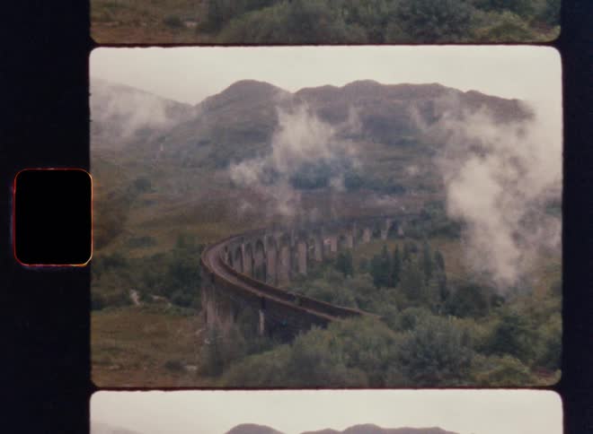 Framed as a film slide a shot shows a steam train crossing a large stone viaduct in a misty green mountain valley
