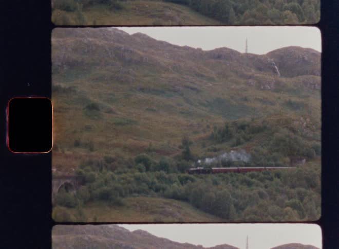 A steam train is moving across a stone bridge through a green hilly landscape with a vintage super 8 film frame look