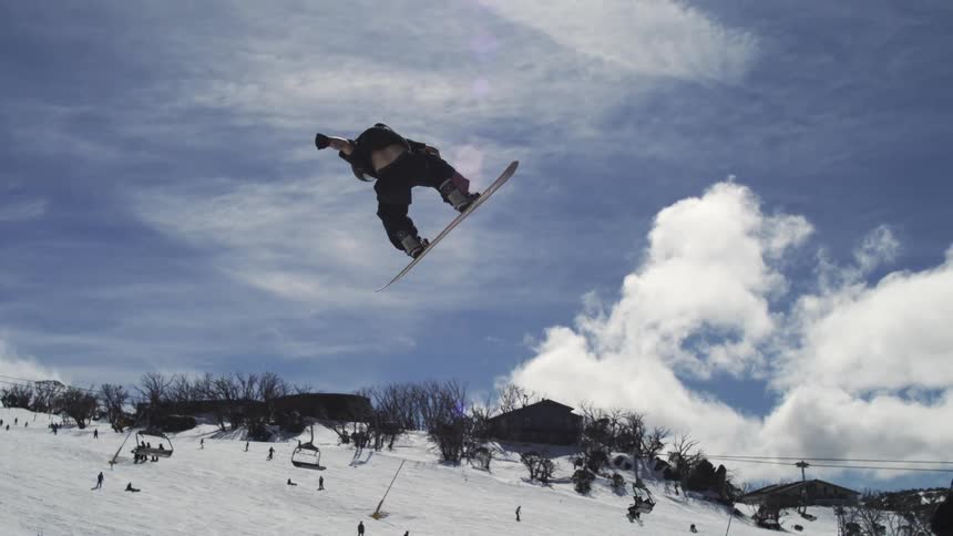 The video shows a person on a snowboard performing a jump high in the air against a bright cloudy sky the person is in a mountainous snowy environment with a ski lift and other skiers in the background a filmsupply watermark is visible in the middle