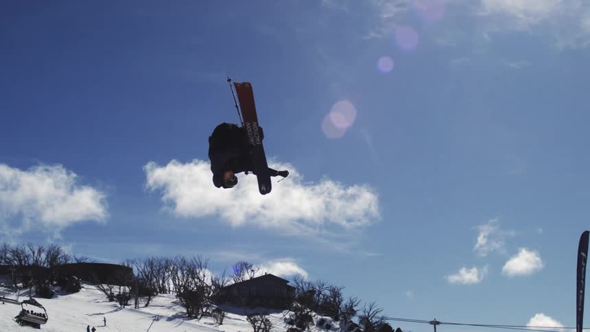 The video shows a freestyle skier in the air performing a flip or a trick the first half of the video captures the jump against the snowy slope and a blue sky with scattered clouds the second half zooms in on the skier whose silhouette is visible agai