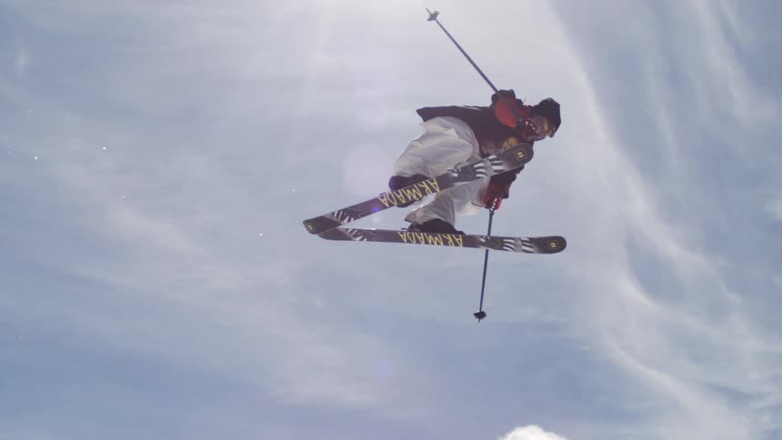 A man is skiing down a snowy slope with his legs straight and his ski poles in his hands with a bright sky in the background