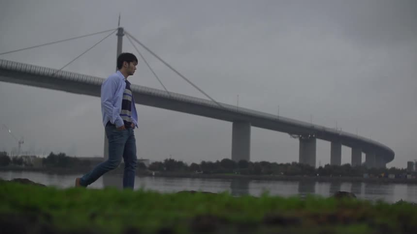 Man walking across the shore with a large bridge behind him