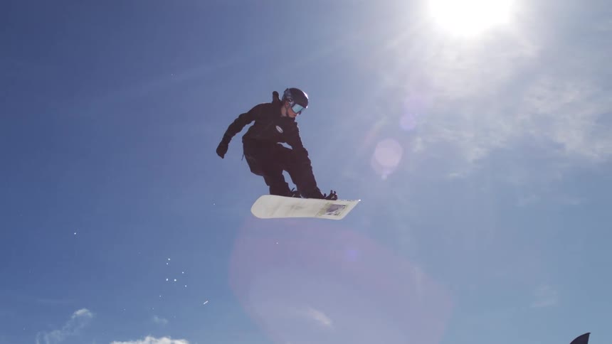 The video shows a snowboarder midair performing a trick against a bright blue sky with scattered clouds the snowboarder is wearing a dark jacket and pants and has their right hand raised in the air the video is set on a snowy mountain with trees and