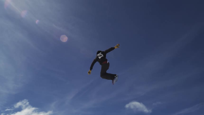 A snowboarder in a dark jacket is performing an acrobatic trick high in the air against a bright blue sky with the sun creating a lens flare and the snowy ground far below